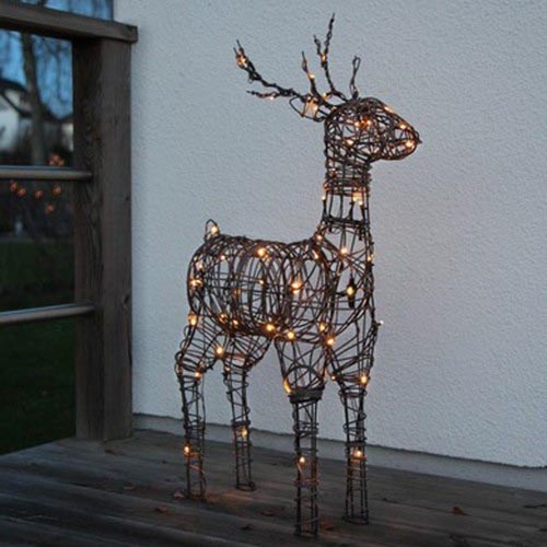 Svietiaca kovová socha jelena stojí na drevenom povrchu terasy pred bielou stenou. Jelen je osvetlený malými teplými svetlami.