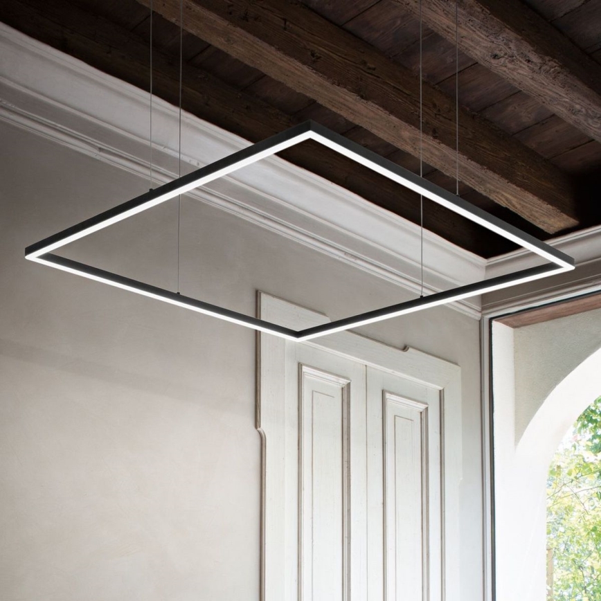 Štvorcová LED lampa visí zo stropu s drevenými trámami. Lampa je umiestnená v interiéri s bielymi stenami a dverami. Vidno časť okna s výhľadom do zeleňe.