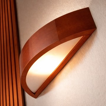 Drevená nástenná lampa v tvare polmesiaca svieti. Svetlo presvitá z mliečne bieleho difúzora. Lampa je z tmavo hnedého dreva.