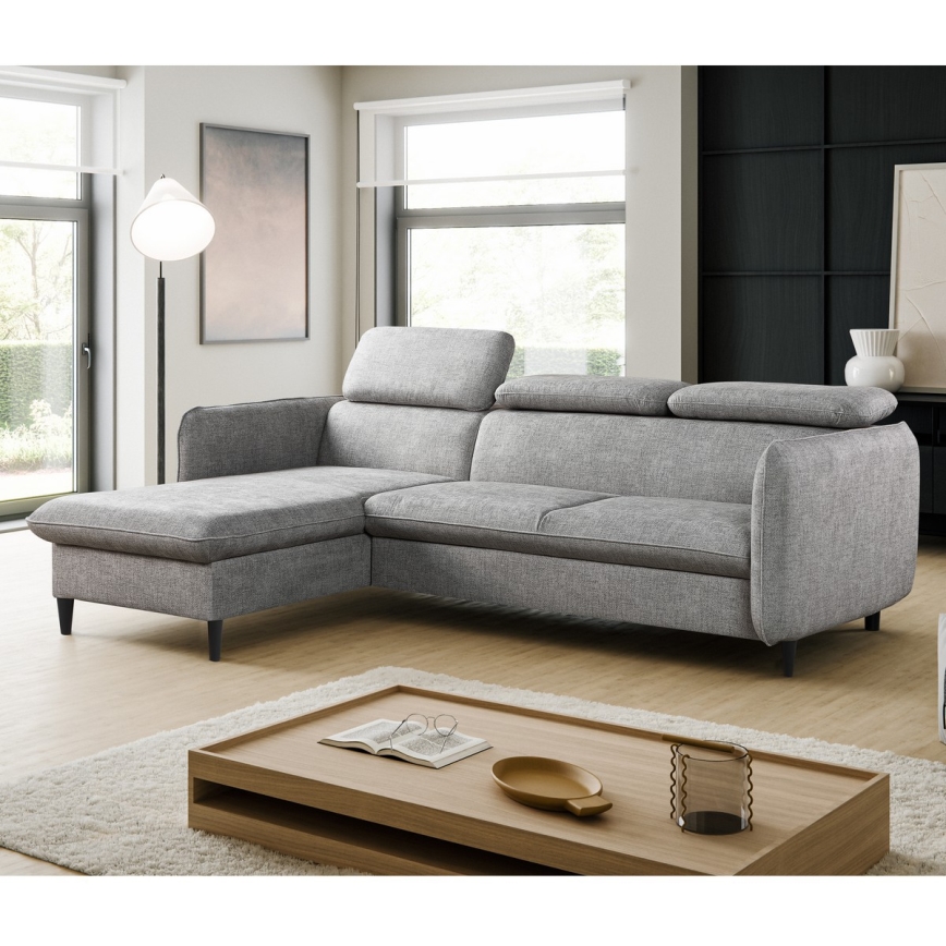 Rohová sedačka stojí na bielom pozadí. Sedačka je sivá, látková, s tmavými nohami a má tri opierky hlavy.