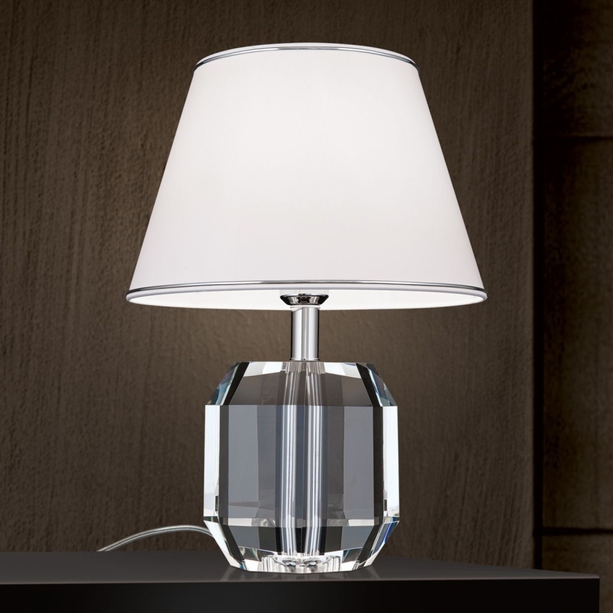 Stolová lampa s kryštálovým podstavcom a bielym tienidlom stojí na tmavom povrchu. Lampa je zapnutá. Tienidlo má tenký kovový lem.