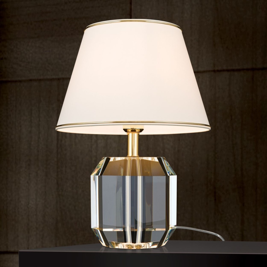 Stolová lampa s kryštálovou základňou a bielym tienidlom so zlatým lemovaním stojí na tmavom povrchu pred tmavohnedou stenou. Lampa je zapnutá.